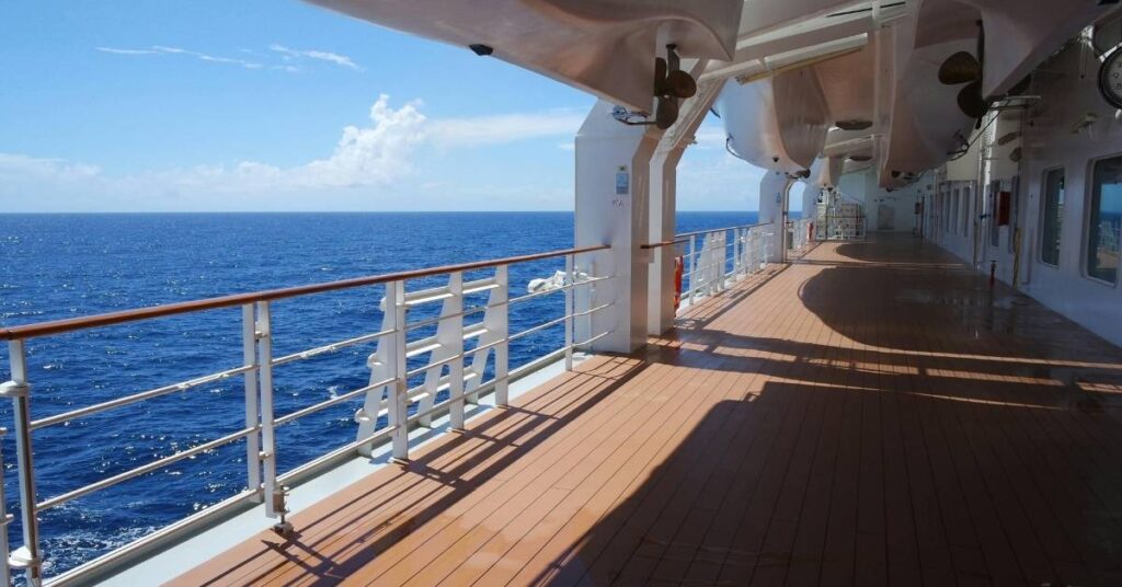 Sunny cruise ship deck with wooden flooring and white railings overlooking a vast, calm ocean.