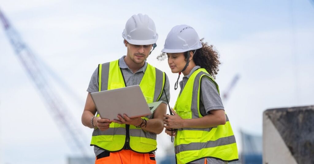 Two individuals in hard hats and safety vests reviewing information on a laptop outdoors.