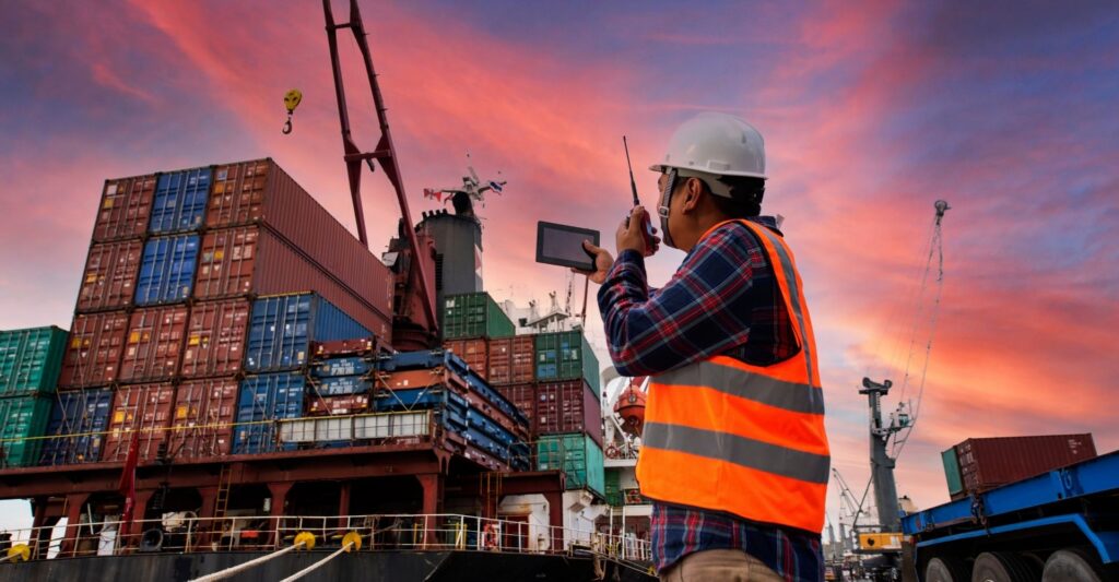 A man in an orange vest stands on a dock surrounded by shipping containers